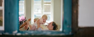 Eine Frau hält ein Baby vor sich hoch. Sie sitzt an einem Tisch in der Küche. Es wurde von draußen durch das offene Fenster fotografiert.