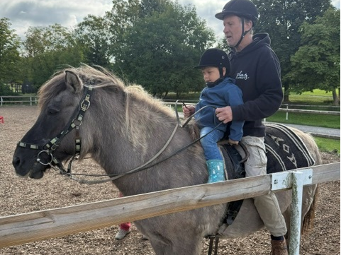 Ein Mann und ein kleines Kind sitzen gemeinsam auf einem grauen Pony. Beide tragen Schutzhelme. Der Mann hält das Kind fest, während das Pony auf einem Reitplatz mit Holzzaun und Bäumen im Hintergrund steht.