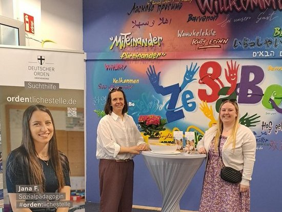 Zwei Frauen stehen lächelnd neben einem Hochtisch mit einem Kuchen und Blumen. Im Hintergrund ist eine bunte Wand mit den Wörtern „Willkommen“ und „Zebra“ zu sehen. Links steht ein Plakat mit einem Porträt einer Frau.