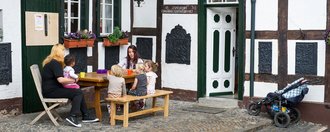 Zwei junge Frauen sitzen mit vier kleinen Kindern an einem Gartentisch vor einem Fachwerkhaus