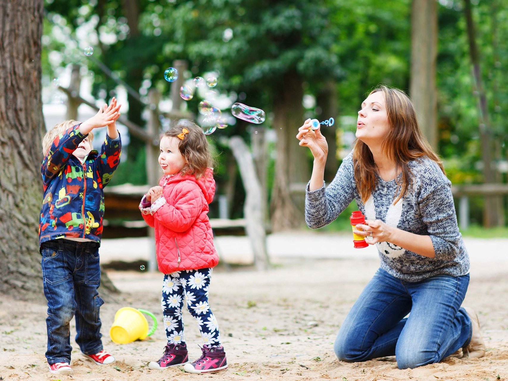 Auf dem Spielplatz: eine Mutter Kniet im Sand und pustet Seifenblasen, die sogleich von zwei kleinen Kindern mit viel Spaß zerklatscht werden 