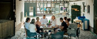 Eine Gruppe Menschen sitzt im Hof eines Bauernhofes um einen Tisch beim Essen.